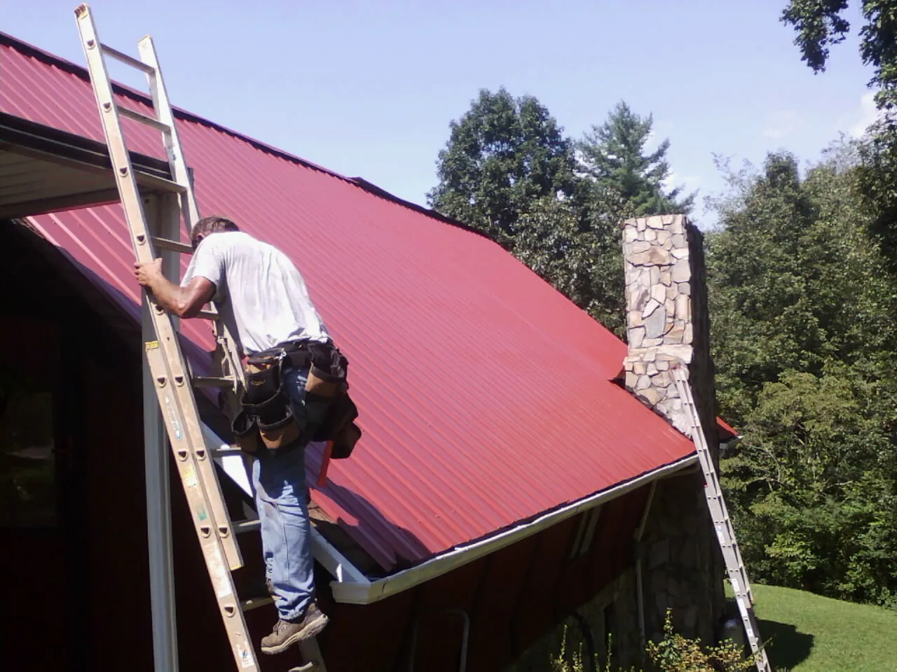 Roofer on a ladder beside a red metal roof and stone chimney — Soffit & Fascia Repair by Roofing Brothers in Burtrum